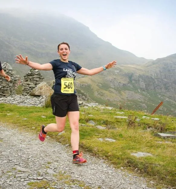 Aimee Jones smiling with open arms while running outdoors on a trail with mountains in the background.