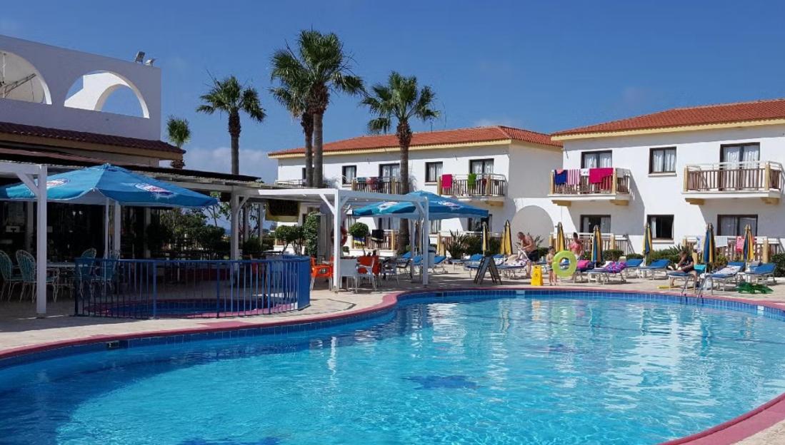 A swimming pool with sun loungers and palm trees, surrounded by white hotel buildings with balconies.