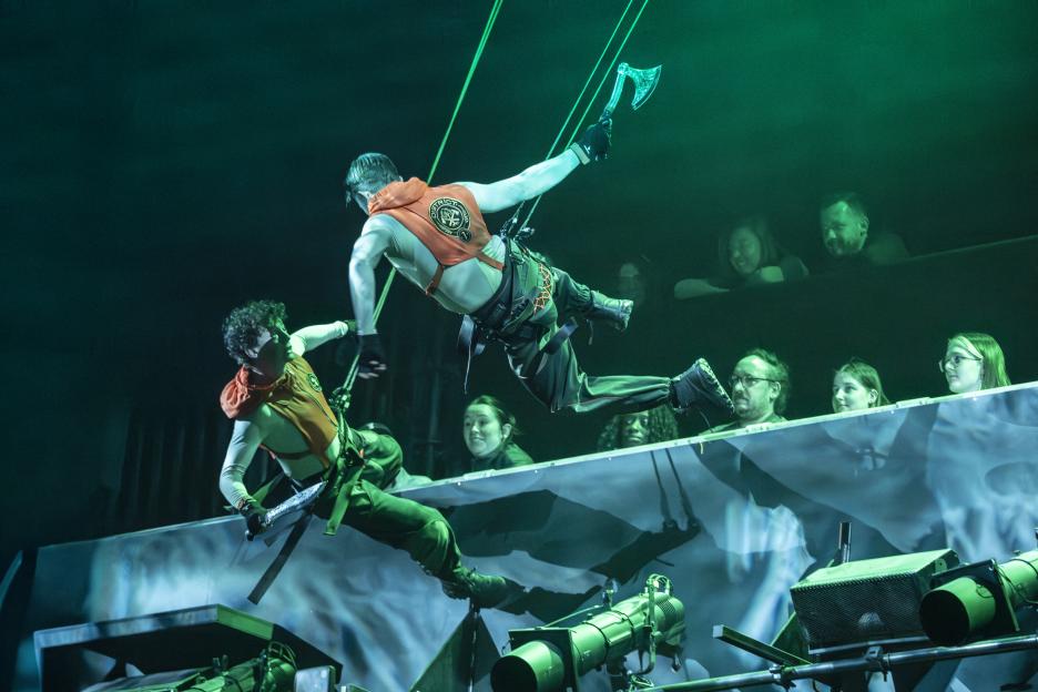 Redmand Rance and Rory Toms performing suspended on stage in "The Hunger Games" play, with Rance holding an axe and Toms a sword.