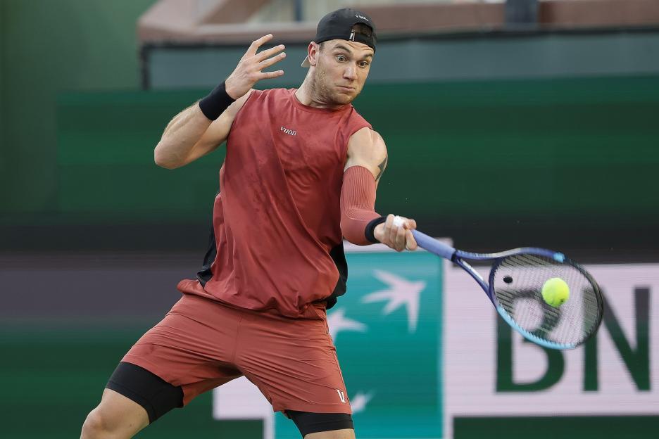 A male tennis player in a red shirt and black cap hitting a forehand.
