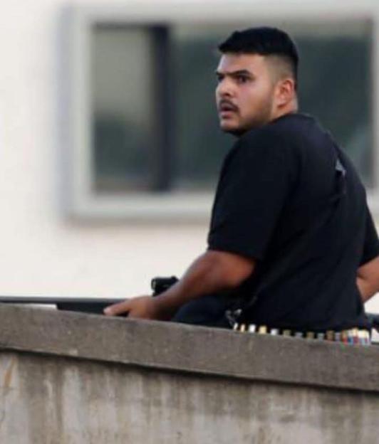 A man in a black shirt and armed with a gun and ammunition, sits atop a concrete wall.