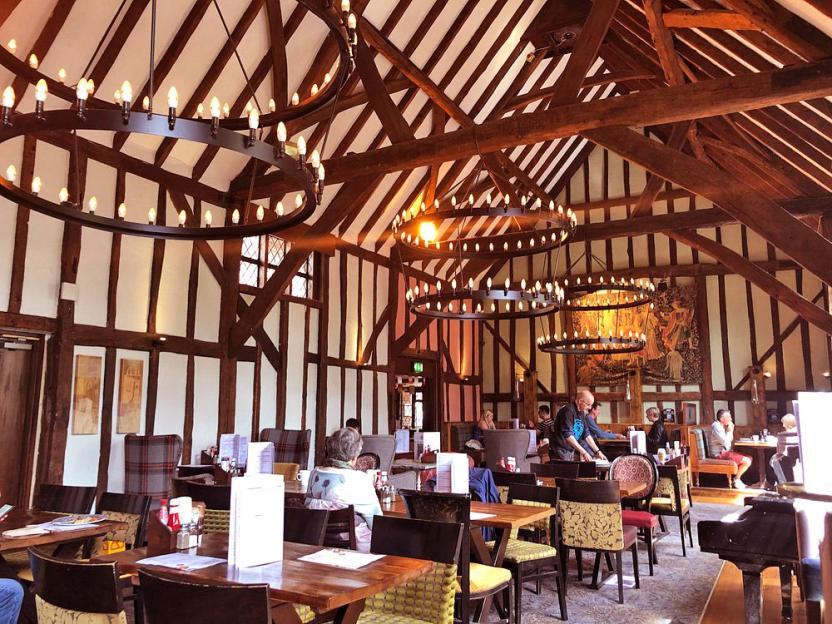 A restaurant with high ceilings, exposed wooden beams, and circular chandeliers, with patrons seated at wooden tables.