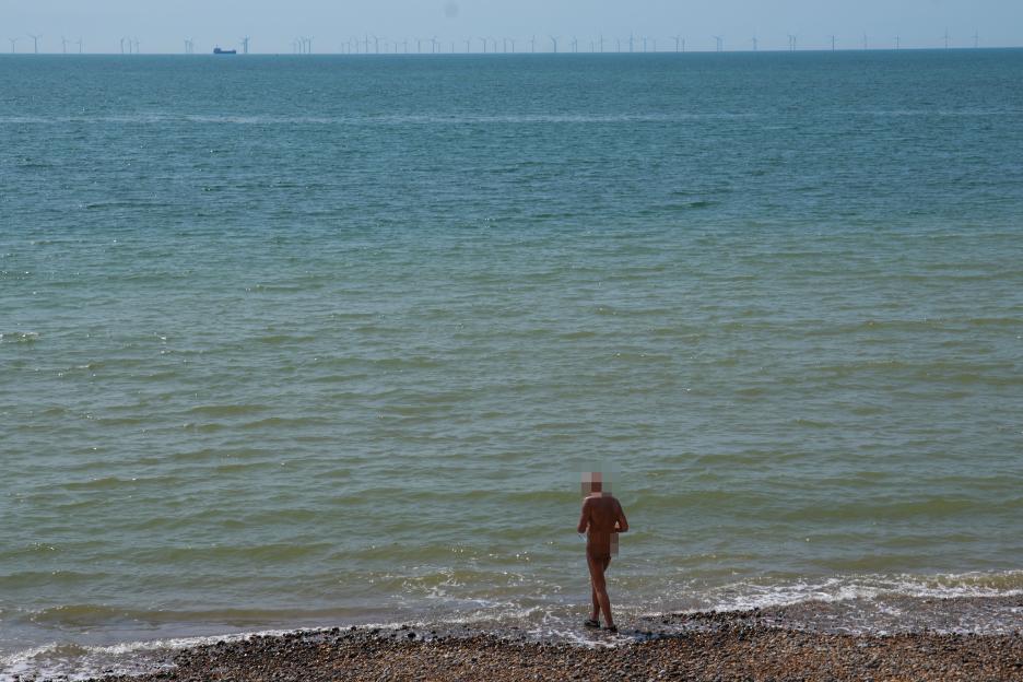 A nude man wading in the ocean at a beach with wind turbines visible in the distance.