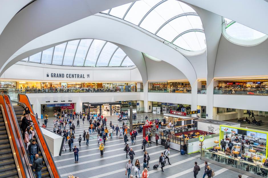 Busy interior of Grand Central Station in Birmingham, England, with people walking on the concourse and escalators, and shops and restaurants visible on multiple levels.
