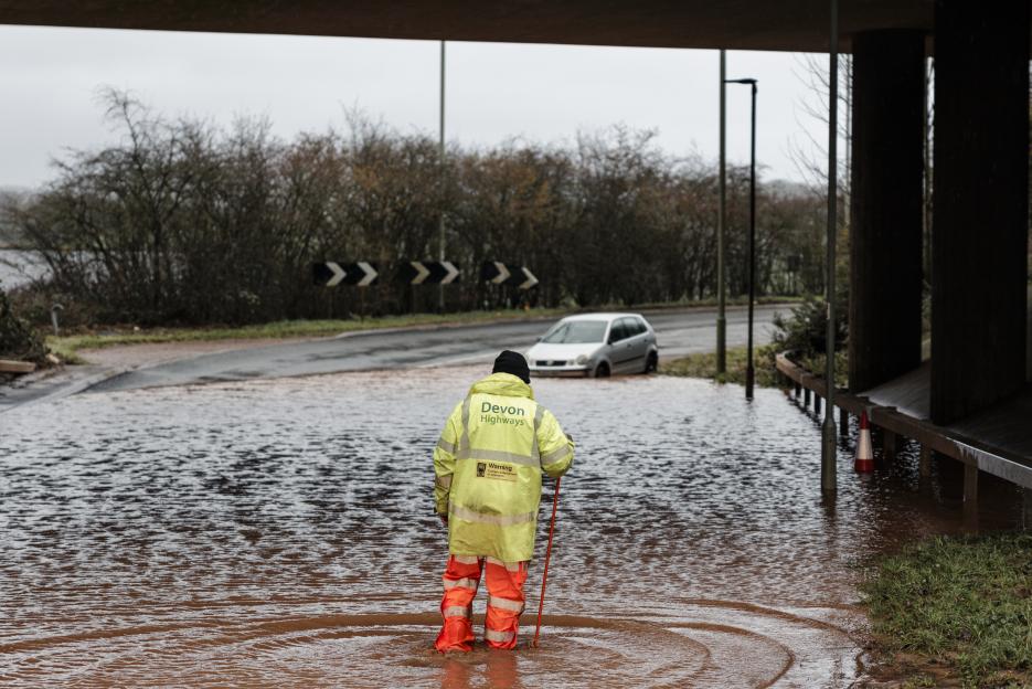 A Devon Highways worker in a high-visibility jacket stands in floodwater next to an abandoned car.