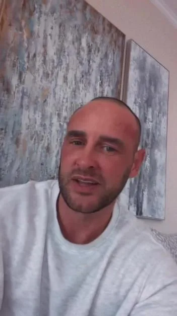 Lee Andrews, wearing a white sweatshirt, speaking in front of abstract paintings.