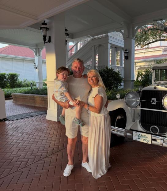 Grandfather holding a child, standing with grandmother in front of a white vintage car.