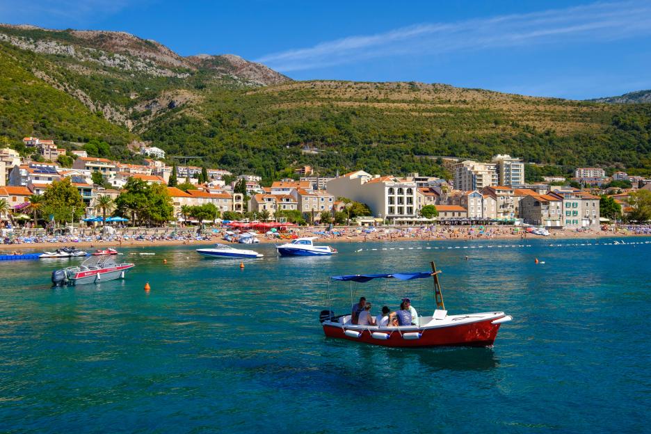 View of Petrovac, a coastal town in Montenegro, with buildings and a beach crowded with people and boats in the water.