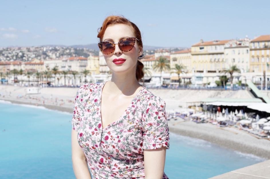 Annette Kellow in sunglasses and a floral dress, overlooking a beach and city.