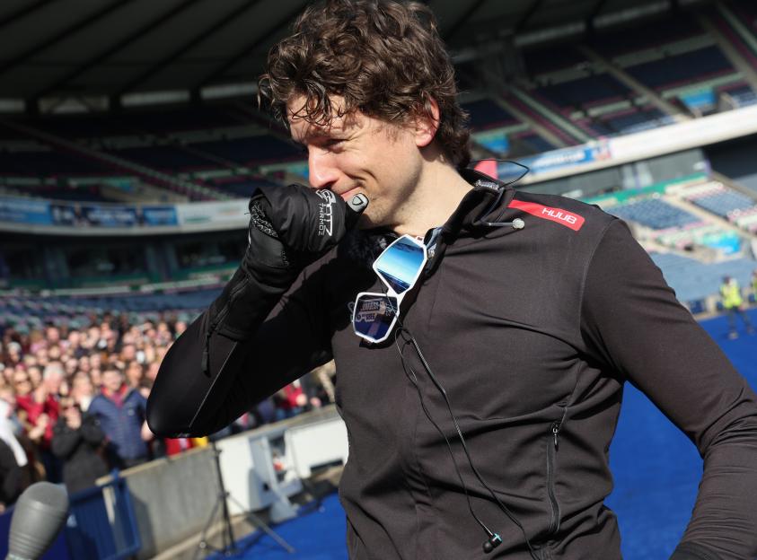 Greg James, sweating with sunglasses hanging from his shirt, touches his nose at the finish line of the Comic Relief bike challenge.