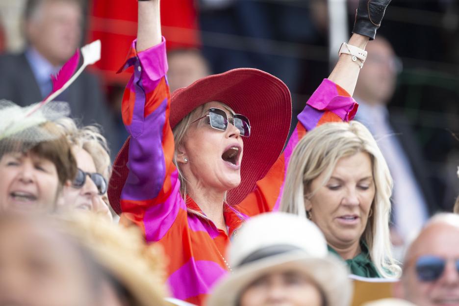 A woman in a red hat and colorful shirt cheers with her arms raised during the Qatar Goodwood Festival.