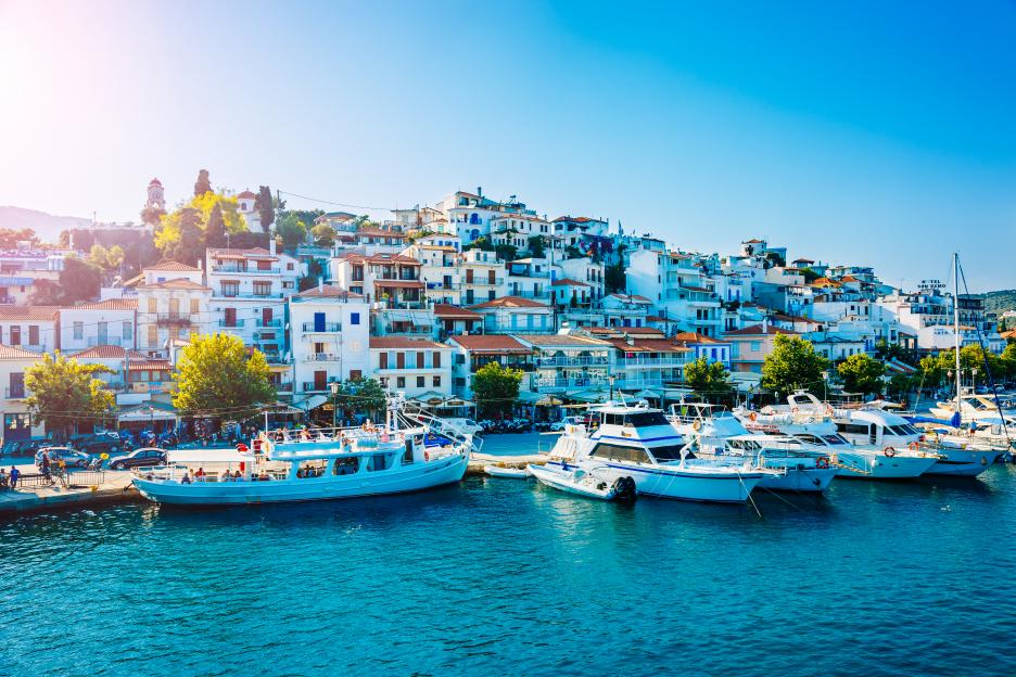 Skiathos town with white houses on a hill, boats docked in the clear blue water, and a bright blue sky.