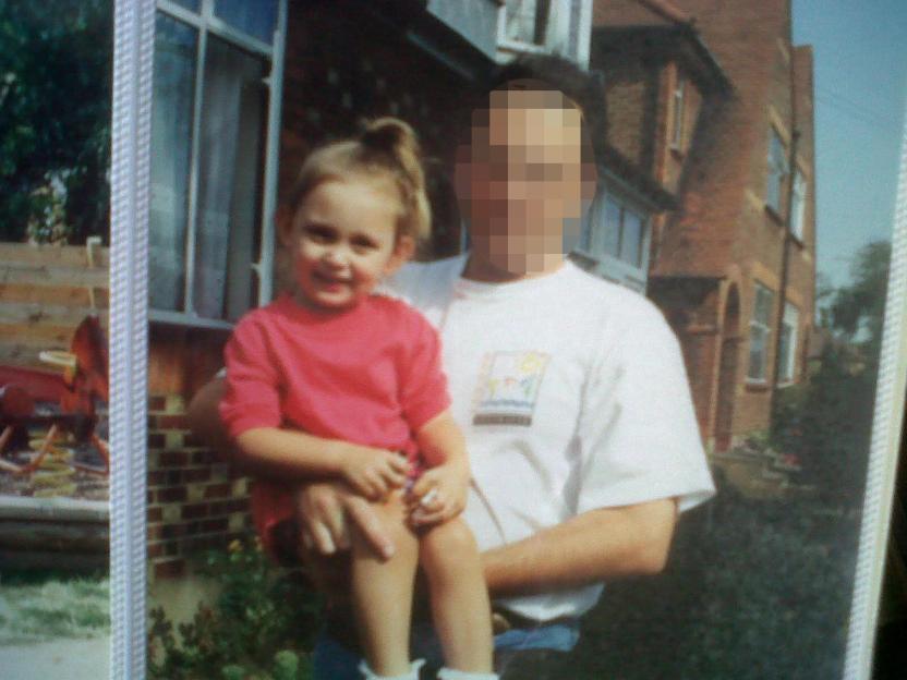 A young girl, Chanel, is held by an adult whose face is pixellated, against a brick house.