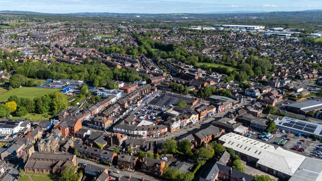 Aerial view of Ashton-in-Makerfield, England, showing residential areas, commercial buildings, and green spaces under a clear sky.