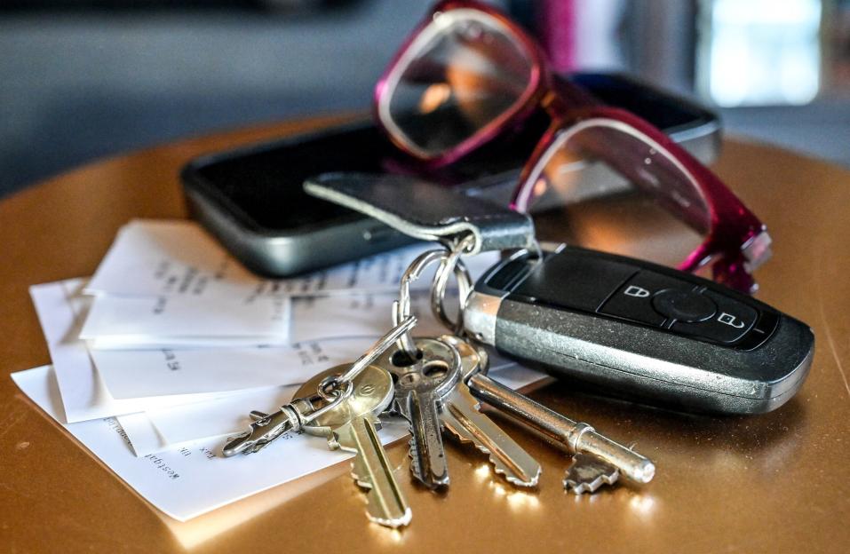 A close-up of keys, a remote car key, and receipts on a table with a phone and glasses blurred in the background.