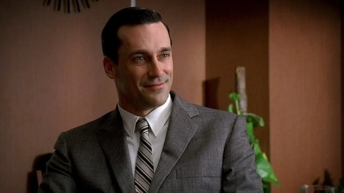 Jon Hamm as Don Draper in a suit and tie, smiling in an interior setting.