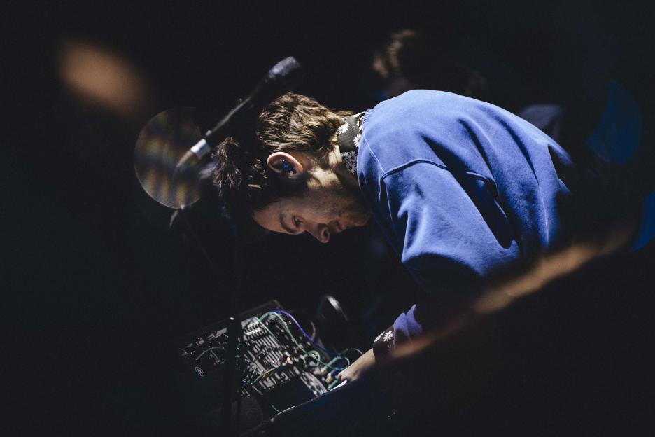 Harry Styles on stage, focused on a music console with wires, wearing a blue sweatshirt and a patterned bandana.