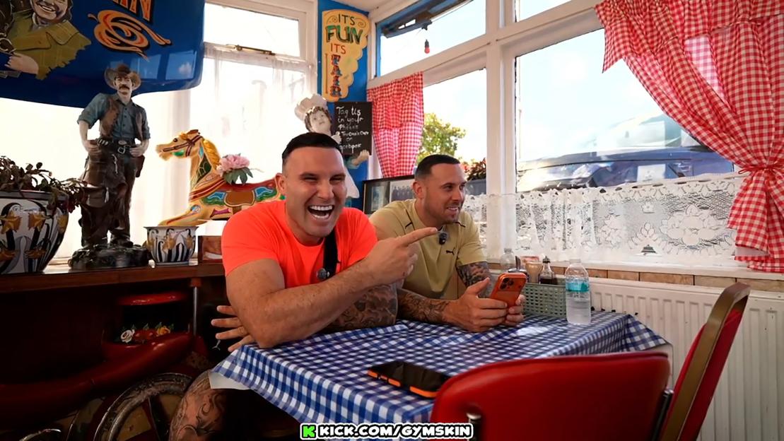 Two men, Jack Smith and Tommy Tokka, at a table in a cafe, one laughing and pointing while the other smiles.