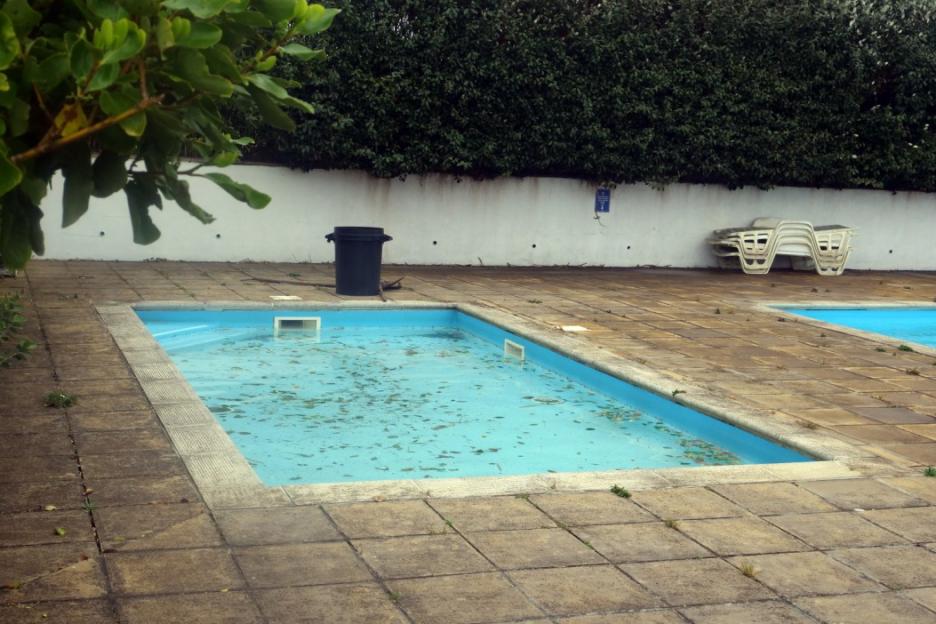 Two abandoned swimming pools filled with leaves, next to a black trash can and stacked white lounge chairs.
