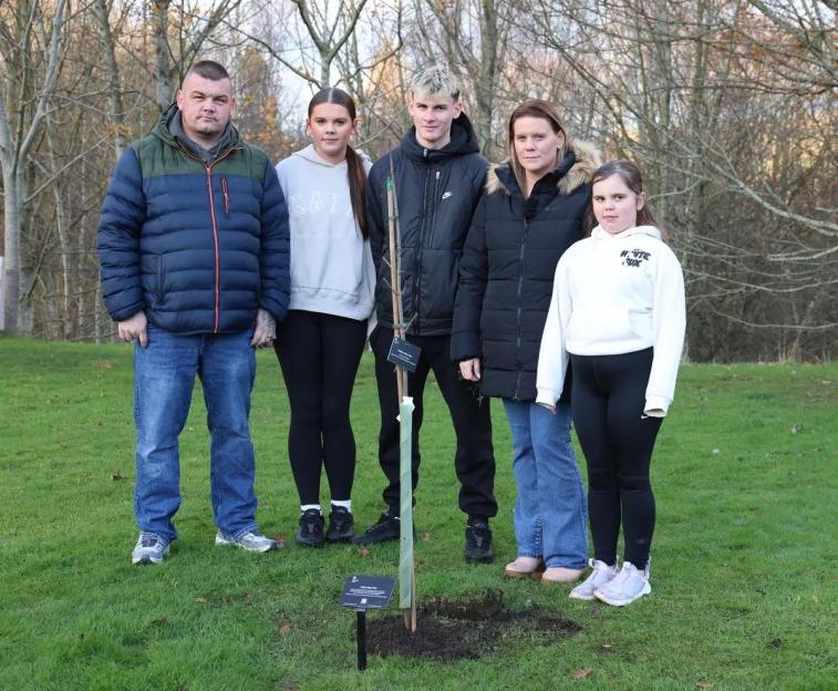 Lee Trussler, two unnamed individuals, Micala Trussler, and another unnamed individual stand in a park next to a newly planted tree.