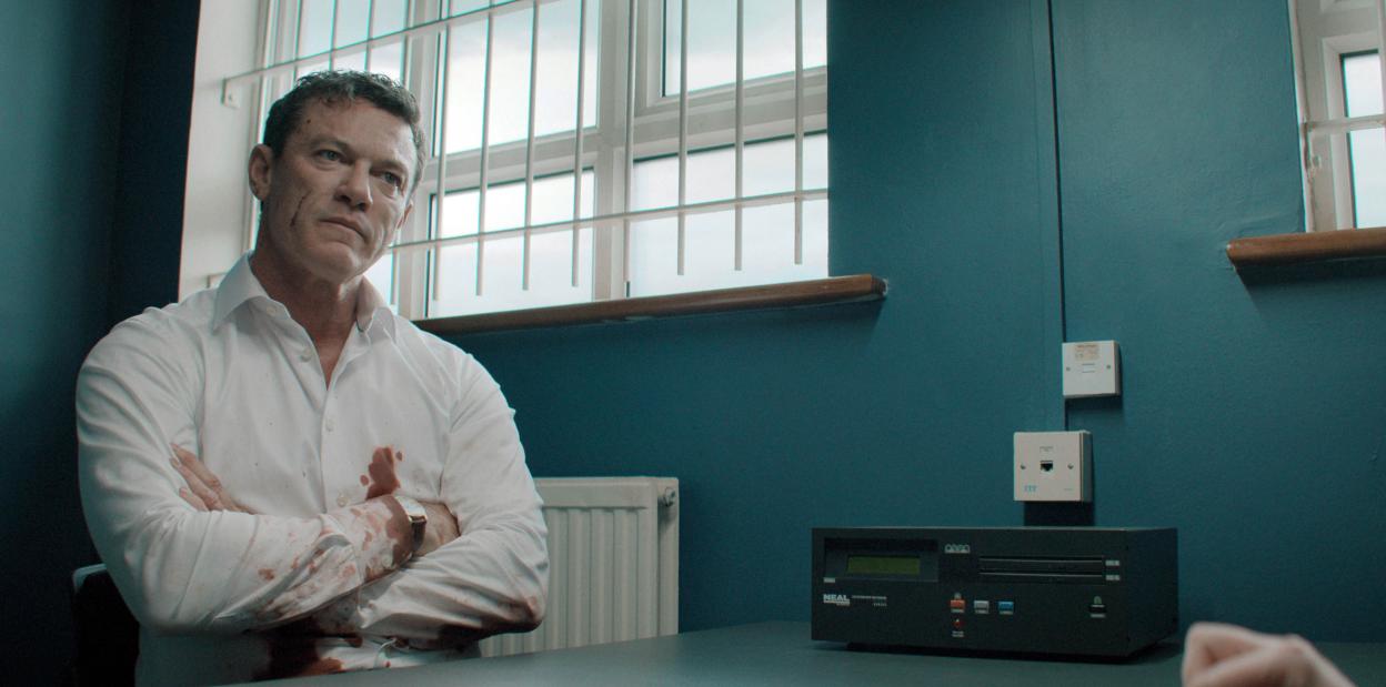 Luke Evans as Martin Gilmour, with a blood-stained shirt, sits in a room with barred windows.