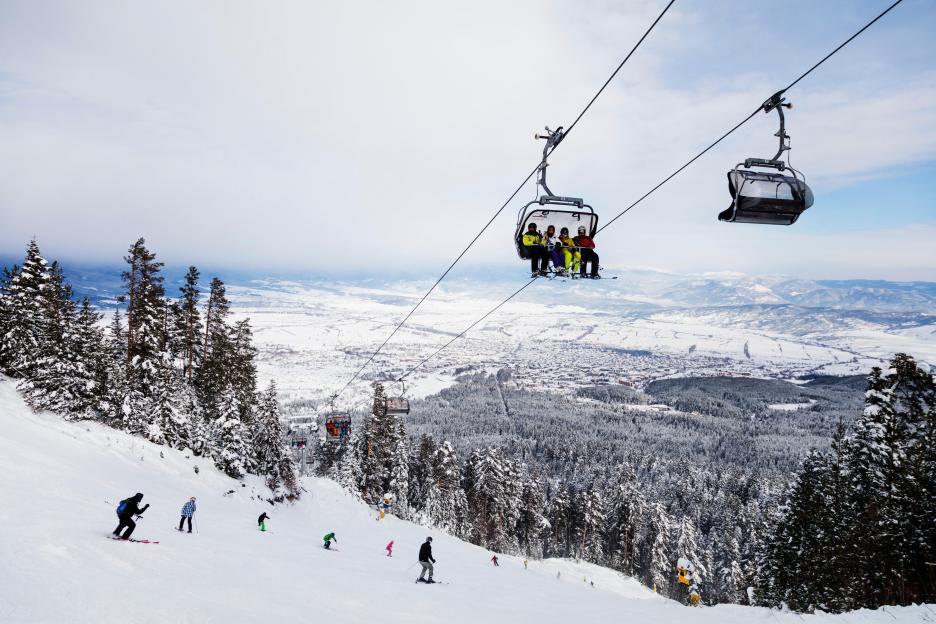 A snowy mountain slope with skiers, pine trees, and ski lifts, overlooking a town and mountains in the distance.