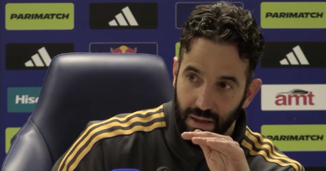 A man with dark, curly hair and a beard speaks at a press conference, wearing a black and yellow striped athletic jacket.