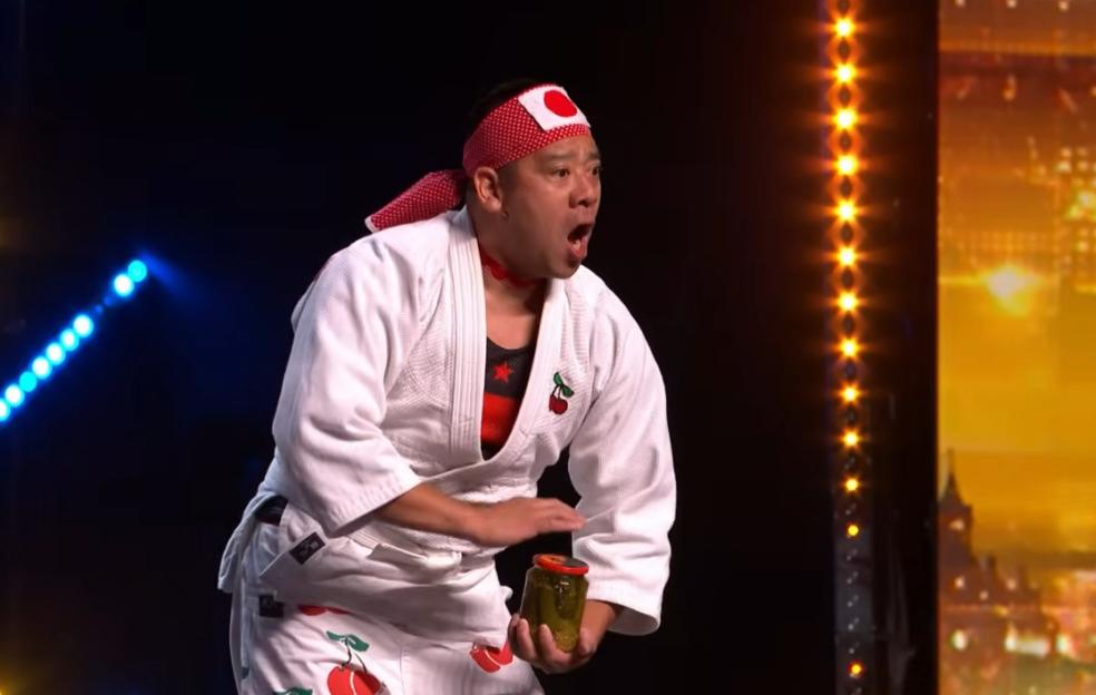 Mr. Cherry from Britain's Got Talent, in a white martial arts uniform with cherry designs, wearing a red and white headband, holds a jar of pickles.