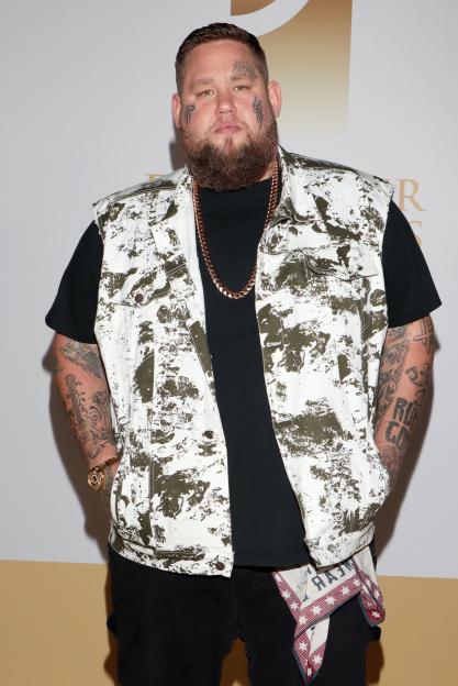 British singer Rory Charles Graham, known as Rag n Bone Man, stands in a black t-shirt and a white and olive green patterned vest with a gold chain.