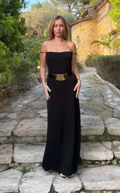A woman in a black off-the-shoulder dress with a wide belt stands on stone steps in a garden.