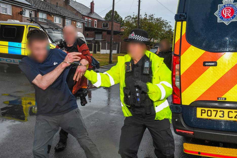 An arrested man in handcuffs being restrained by a police officer from Greater Manchester Police, with a police van in the background.