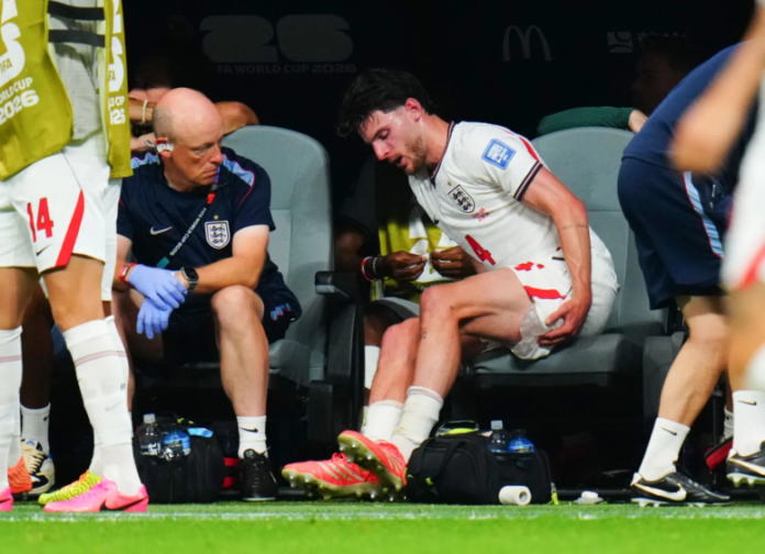 A soccer player in a white uniform with the number 4 applies an ice pack to his thigh while sitting on the bench next to a coach.