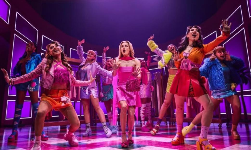 Legally Blonde the musical cast on stage, with a woman in a pink dress and clutch at the center, surrounded by other cast members in colorful outfits.