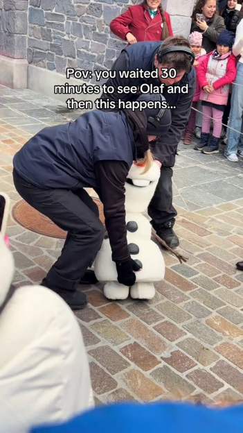 A Disney employee attempts to fix a fallen Olaf robot on a cobblestone path while children watch.