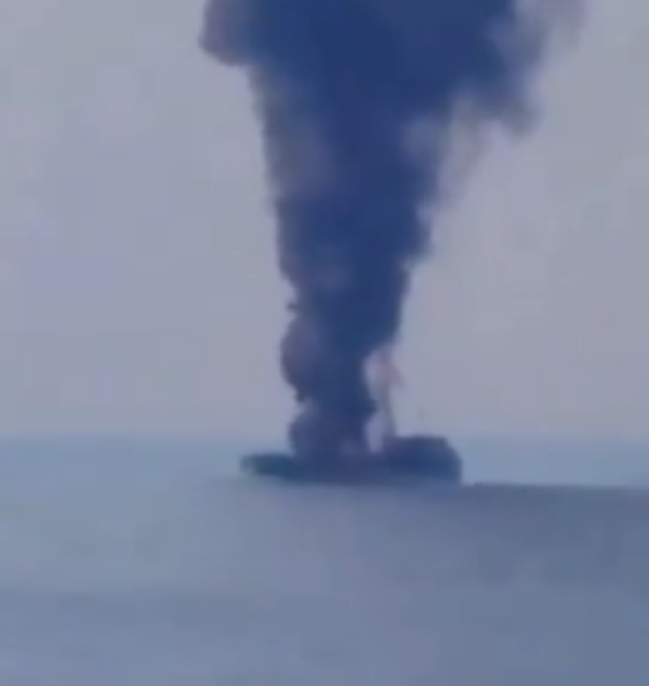 Thick black smoke rising from a burning ship in the water.