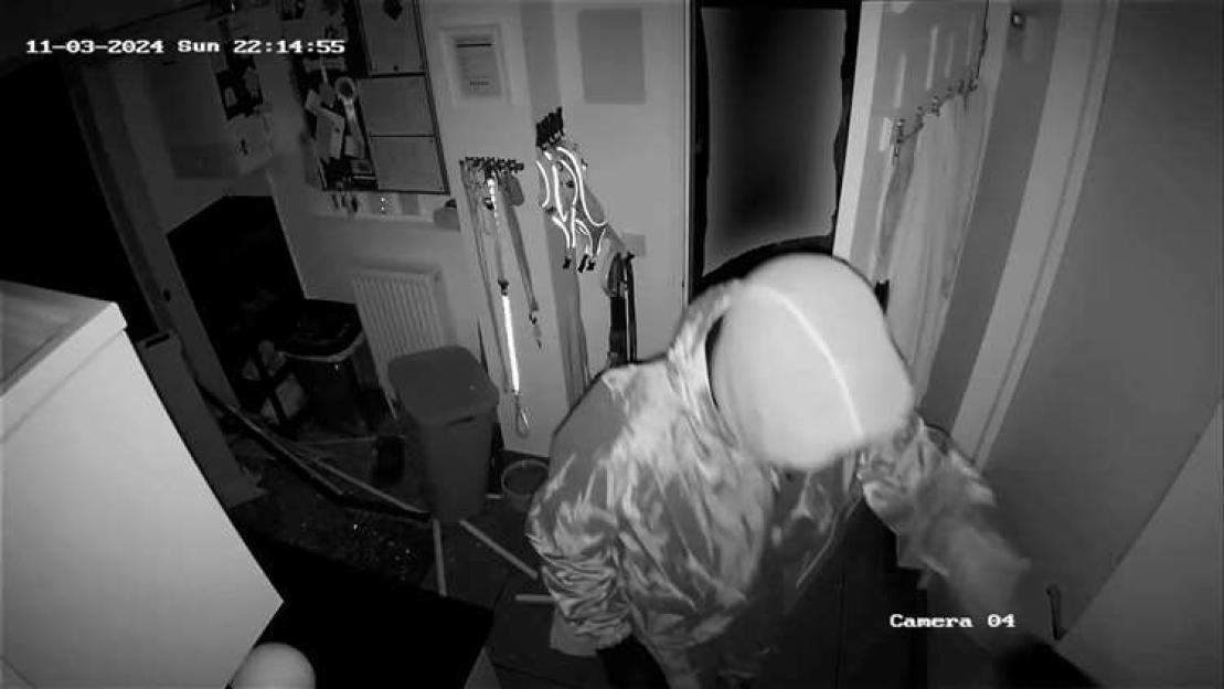 Security camera footage of a person with a covered face wearing a light-colored jacket, entering a room.
