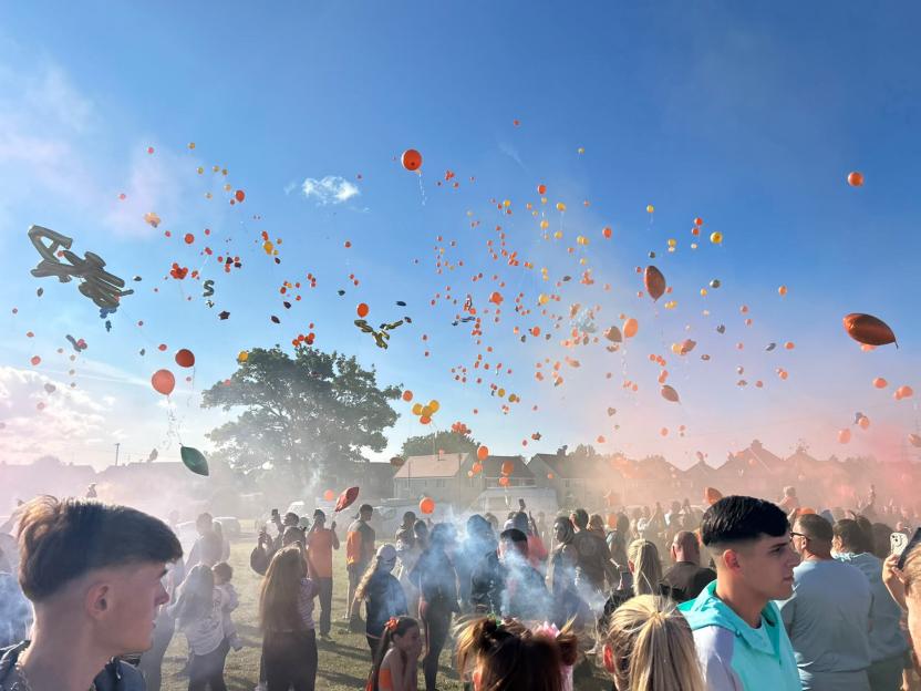 A crowd of people releasing orange and yellow balloons into a blue sky.