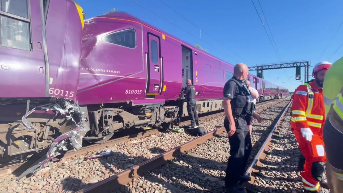 A purple train derailed on train tracks, with emergency personnel in yellow vests on and around the train.