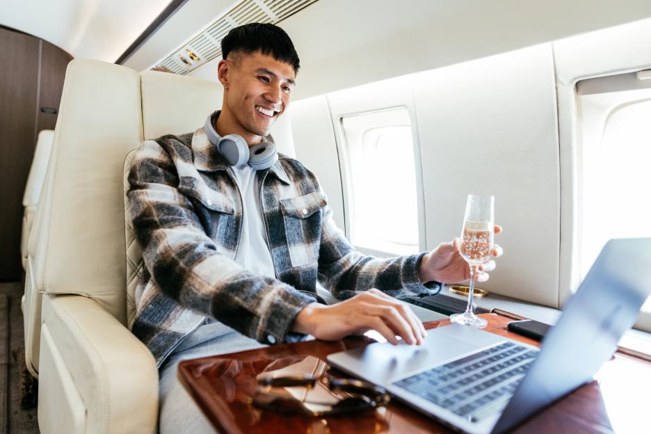 A man smiling while working on a laptop in a private jet, holding a glass of champagne.