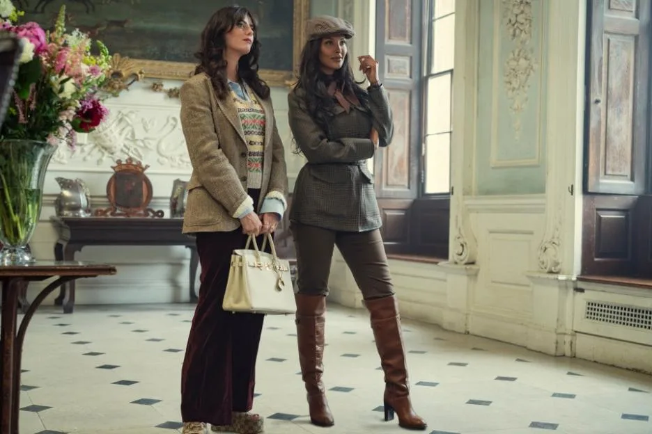 Two women in vintage-style country attire stand in an elegant, sunlit room with a tiled floor, one holding a cream handbag and the other wearing a cap and tall boots.