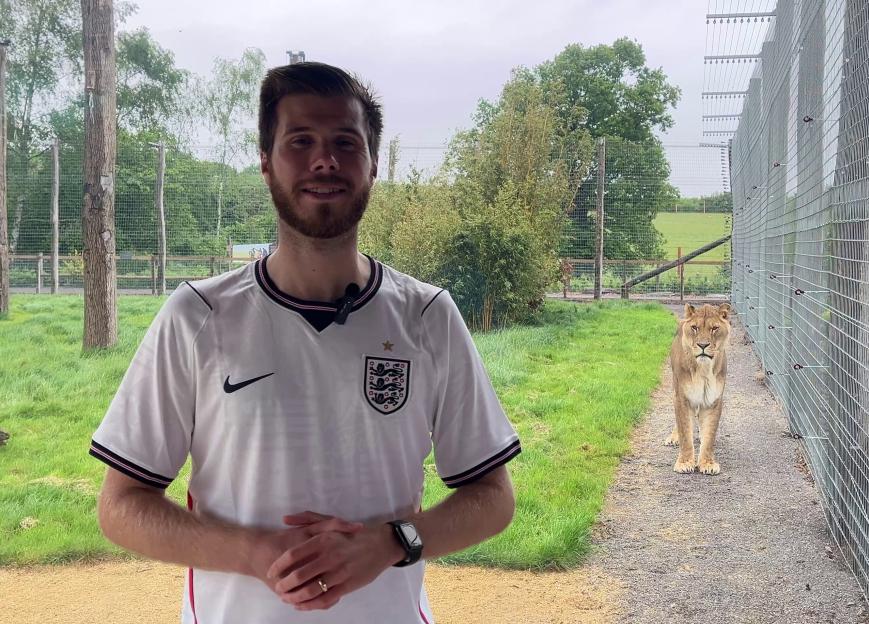 A man in a white jersey stands in front of a chainlink fence, with a lion on the other side of the fence.
