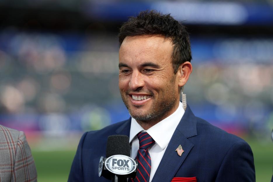 Sports commentator wearing a navy suit, red and blue striped tie, and a US flag lapel pin, smiling and holding a Fox Sports microphone.