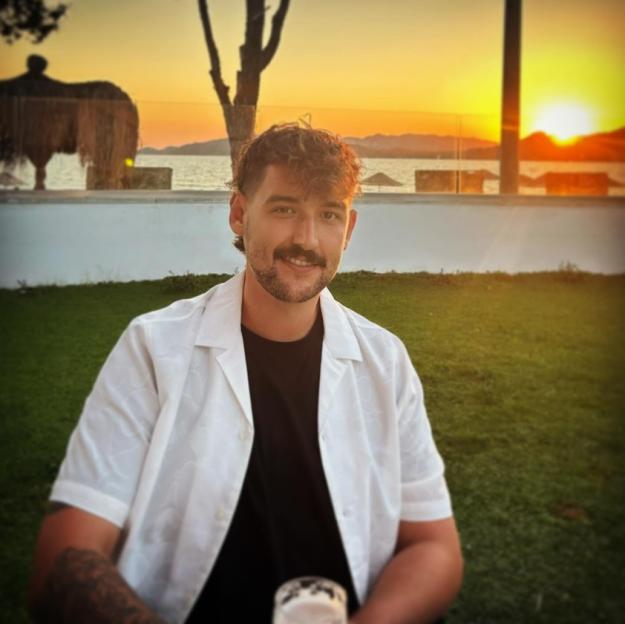 A man with a mustache smiles at the camera with a sunset over water in the background.