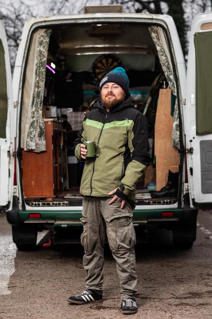 A man in a beanie and winter jacket holding a mug, standing in front of his converted minibus.