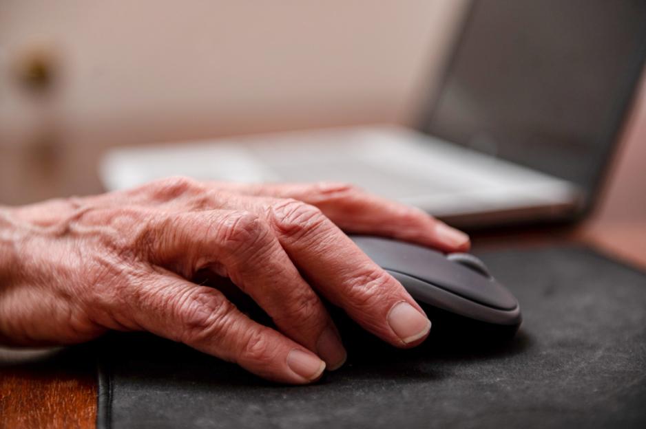 A wrinkled hand on a computer mouse, with a laptop in the background.