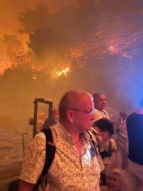 People stand in front of a large fire with orange smoke and embers in the background.