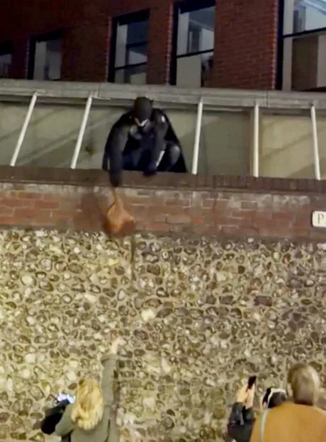Person dressed as Batman on a ledge hands down a bag to people below.