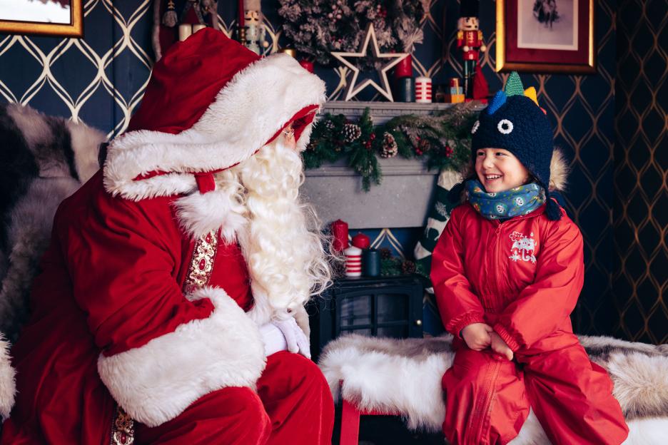 Santa Claus sitting with a child in a red snowsuit and a dinosaur hat.