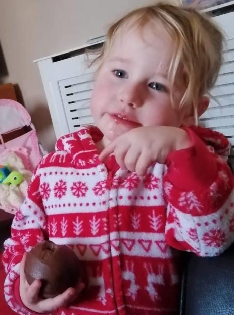 Lola James, a two-year-old girl, wearing a red and white patterned onesie, holds a chocolate egg.