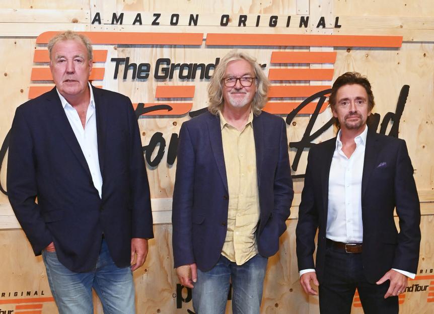 Jeremy Clarkson, James May, and Richard Hammond posing in front of a sign for "The Grand Tour."